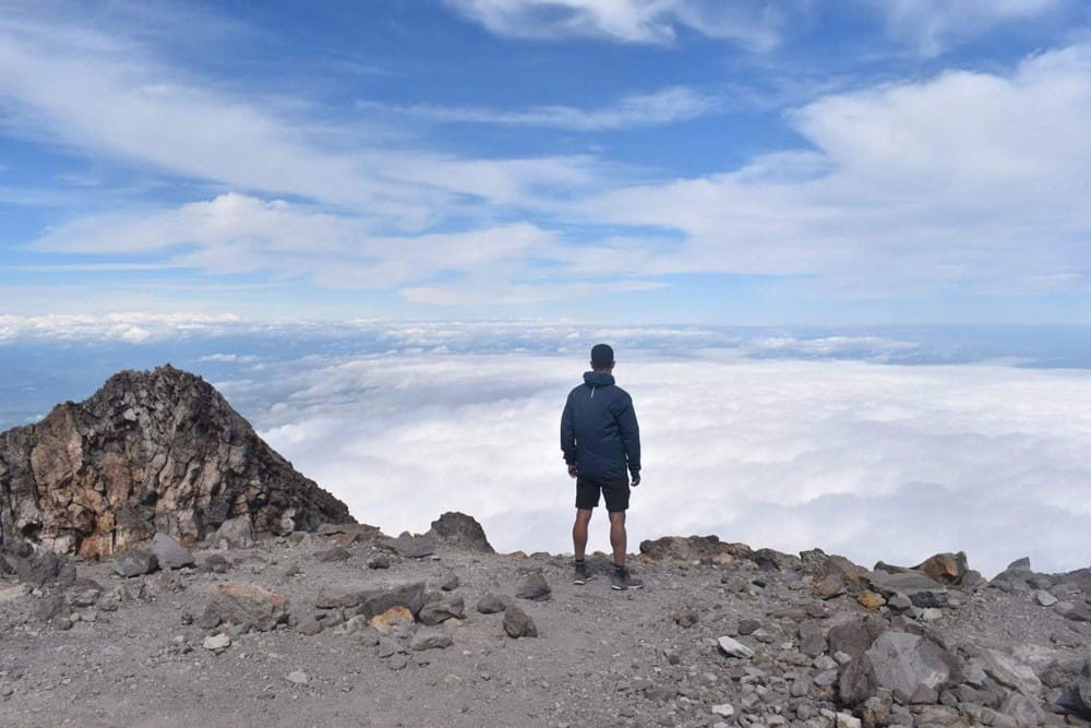 Lugares de Nueva Zelanda chico moreno de espaldas en la cima de una montaña y delante las nubes
